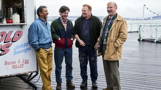 The cast of Men Up stand on a wet and rainy seaside pier in Wales next to a hut selling "jumbo sausages" (geddit?) 