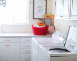 A laundry room with a top load washing machine, white cabinets and red plastic bucket filled with cleaning supplies