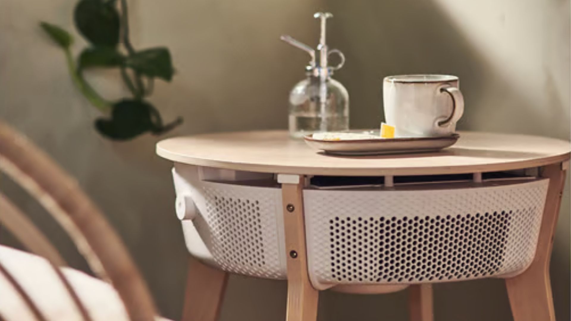 The image shows a pale wooden bedside table in a neutrally decorated bedroom, with a coffee cup and saucer on top. Beneath the table is a white integrated air purifying system.