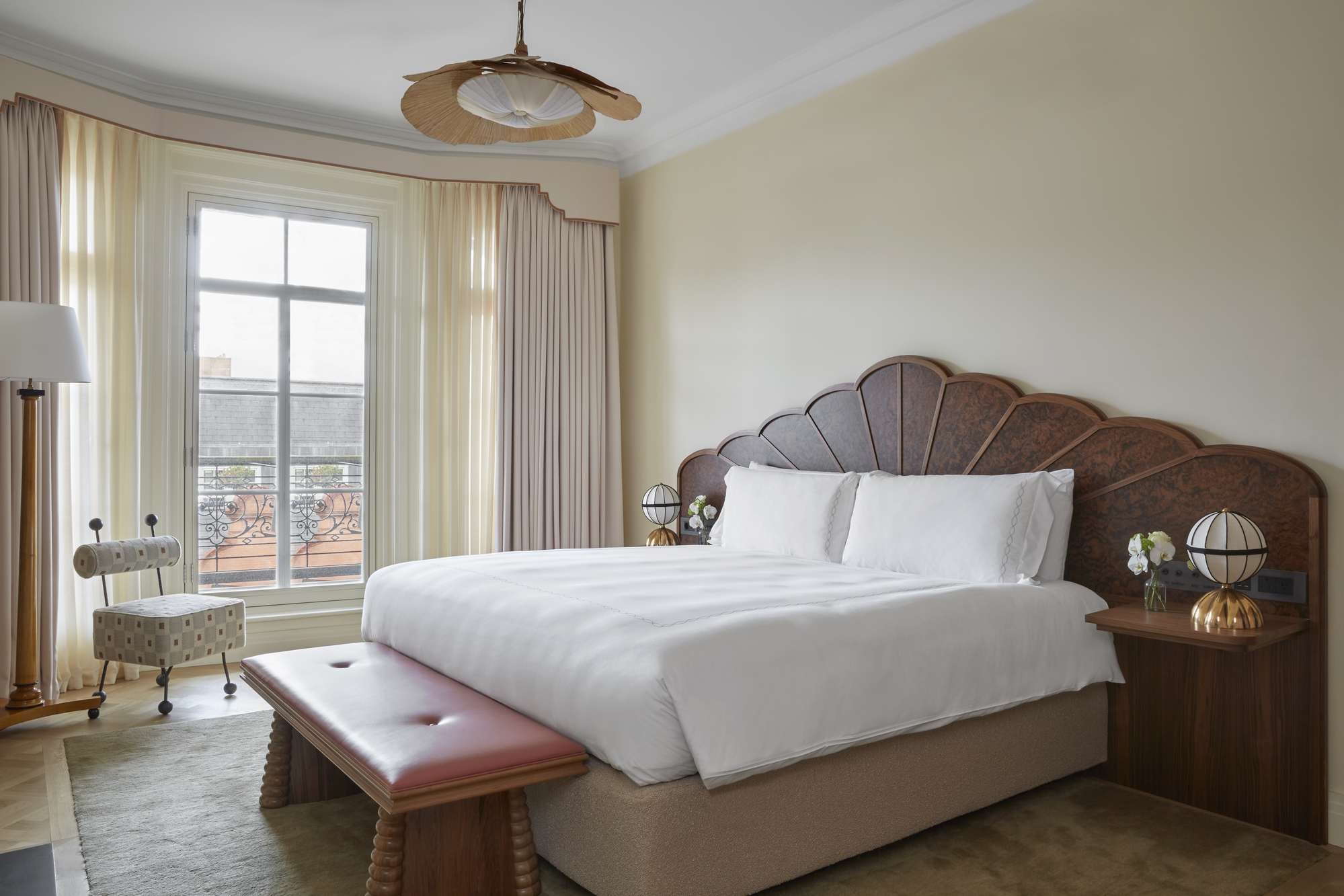 Image of a hotel bedroom at London's Claridge's hotel. The room is a pale yellow with a large window that has beige curtains. The bed has an extended wooden headboard with a fanned design, and the bedding is all white. There is a red leather bench seat in front of the bed.