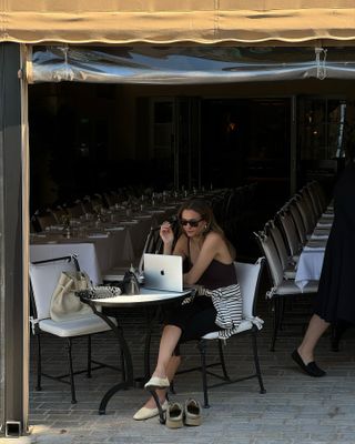 Anne-Laure Mais sitting at a cafe on her laptop wearing a camisole and capri pants with flats and a striped shirt around her waist.