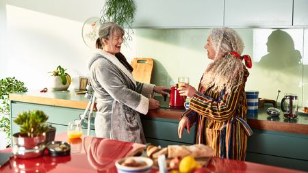 Two female senior roommates stand chatting in a kitchen over breakfast.