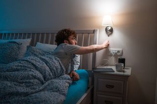 A young Caucasian male lying in bed, unable to sleep, reaching out to a bedside lamp on darker morning..