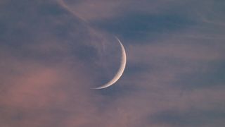  A thin crescent moon is pictured against a darkening blue sky at sunset with a sliver of its right side lit by direct sunlight. Wispy clouds are visible in the foreground. 