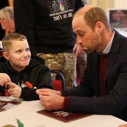 Prince William sitting at a table looking at a paper with a pouting face next to a little boy