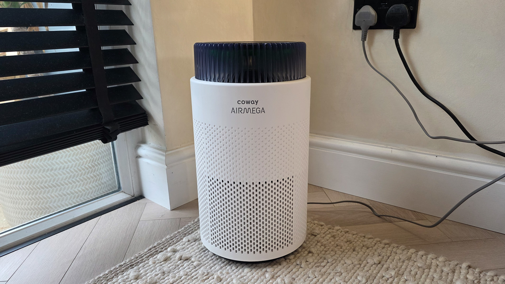 The Coway Airmega 100 standing in the corner of a room near some blinds.