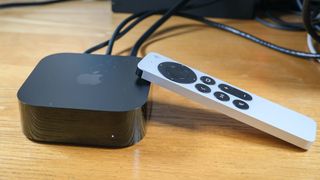 The Apple TV 4K (2022) is on a wooden surface, with its remote propped against it and cords coming out of the back.