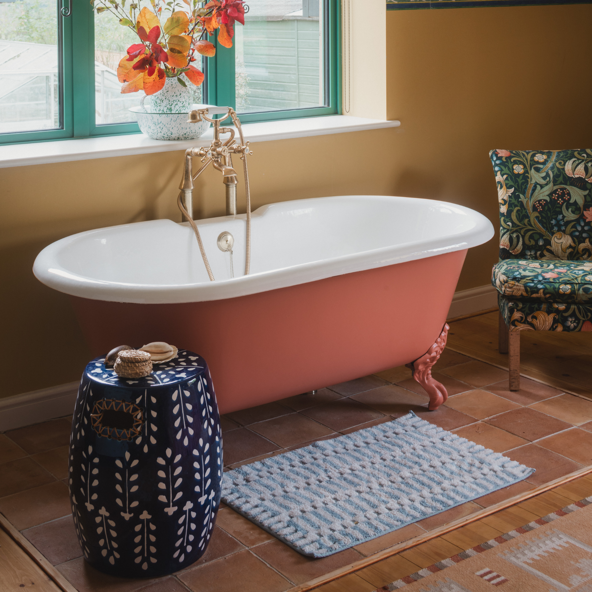 Terracotta bath in yellow and terracotta bathroom with blue side table and green chair and window panes.