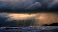 A dramatic photo of a rain storm over the ocean with orange sunlight peeking through dark clouds