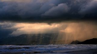 A dramatic photo of a rain storm over the ocean with orange sunlight peeking through dark clouds