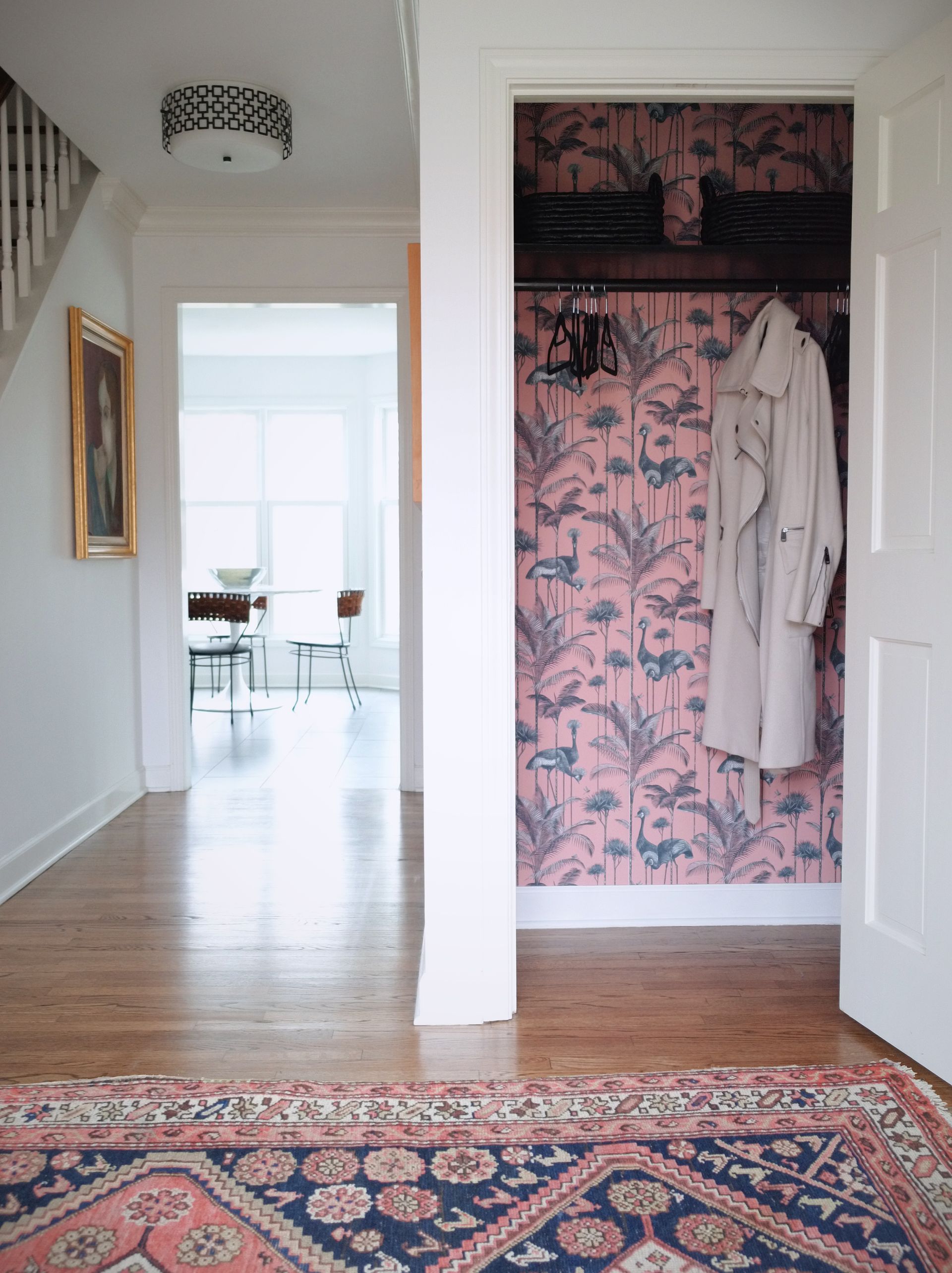 closet with black hanging rail, black baskets and hangers, mac hanging up, crane wallpaper behind, entrance way, rug, hardwood floor, dining table in background and stairs