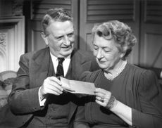 An older couple from the 1950s looks at a check and smiles. The photo is in black and white.