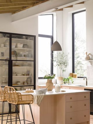 pink and white kitchen with pink units and island, black crittall style window frames and black framed glass fronted pantry, gray pendant light, rattan bar stools