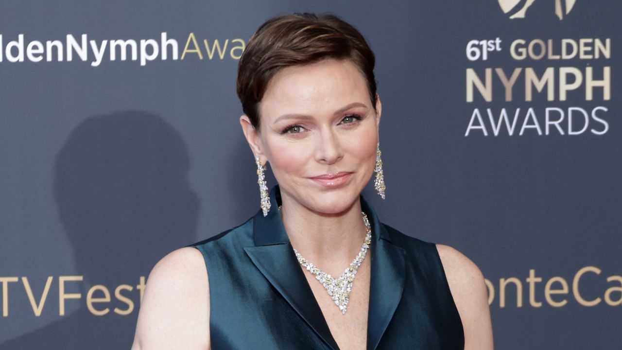 Princess Charlene of Monaco wears a black shirt dress with a large diamond necklace and matching drop earrings while her hair is brunette and cropped short