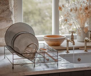 A set of speckle glaze plates drying on a small metal dish drying rack, next to a set in kitchen sink with gold faucets. In front of a kitchen window set into a stone wall, with dried flowers in a vase.