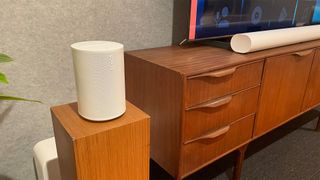 Sonos Era 100 wireless speaker sitting on wooden shelf in living room setting with TV and Sonos Arc 2 soundbar