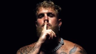Jake Paul putting his finger to his lips to shush someone. He's shirtless and wearing a silver chain. The room is dark around him. 