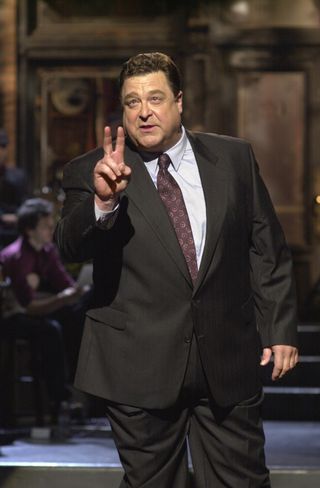 John Goodman in a suit holding up two fingers while hosting snl