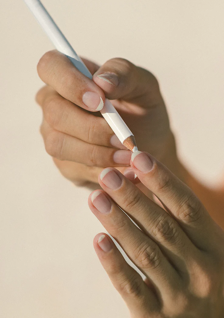 two hands on a nude background with bare nails and one hand coloring underneath the tip of a nail with a white pencil