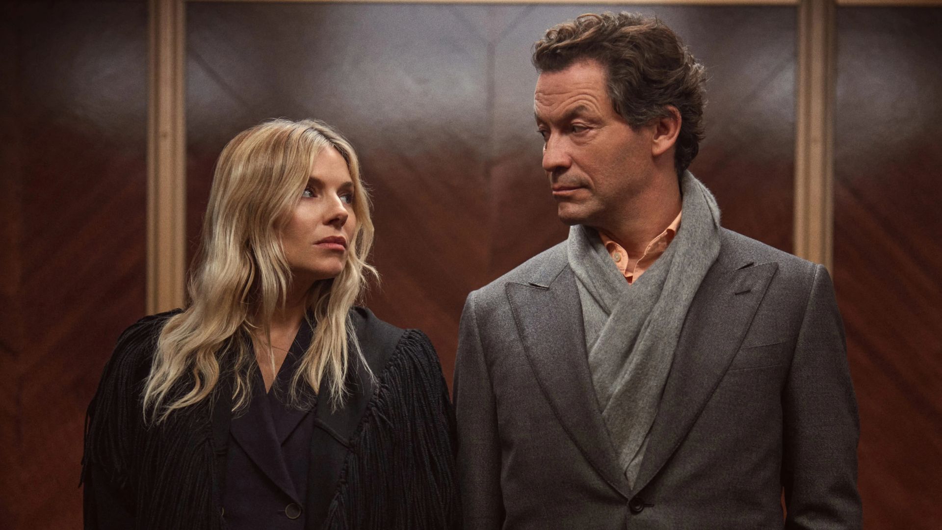 Sienna Miller and Dominic West look at each other in an elevator