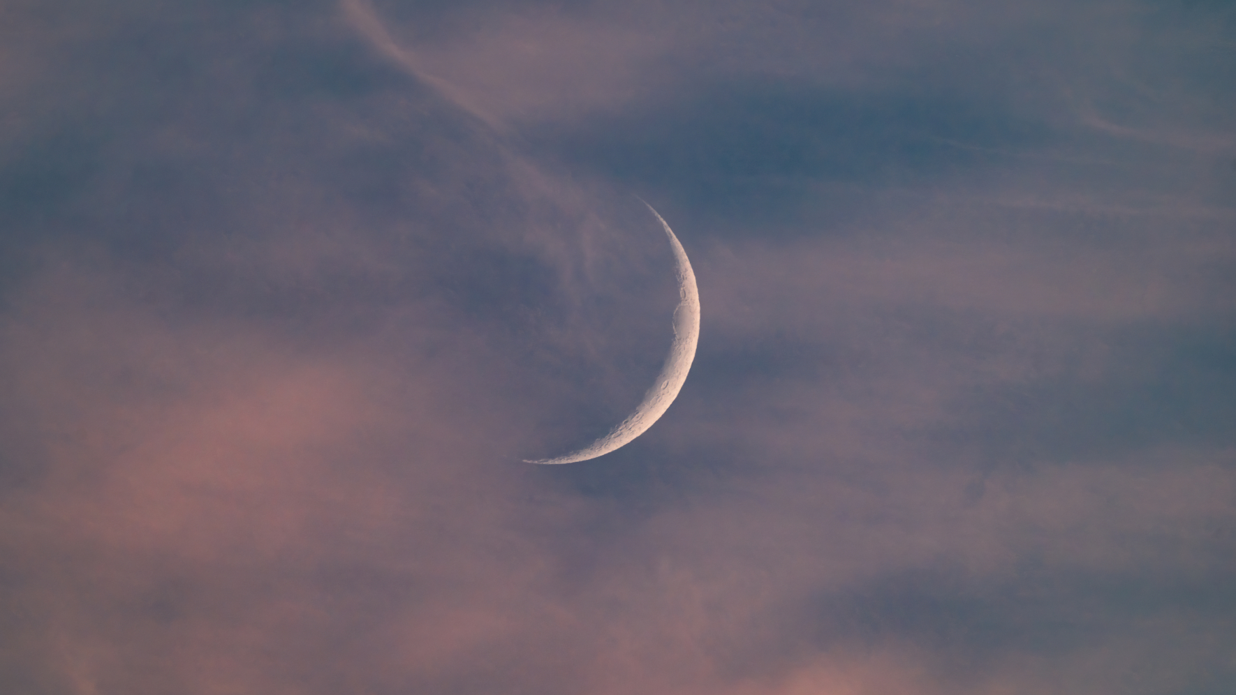 A crescent moon is pictured in a cloudy evening sky close to sunset.