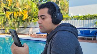 The reviewer wearing the Yamaha YH-L700A headphones and looking at a smartphone. In the background are a swimming pool and palm trees.