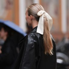 A guest at Copenhagen Fashion Week wearing silver earrings, cream white satin and silk ribbon as hair accessory, black leather long coat with a slicked ponytail and the best hairspray