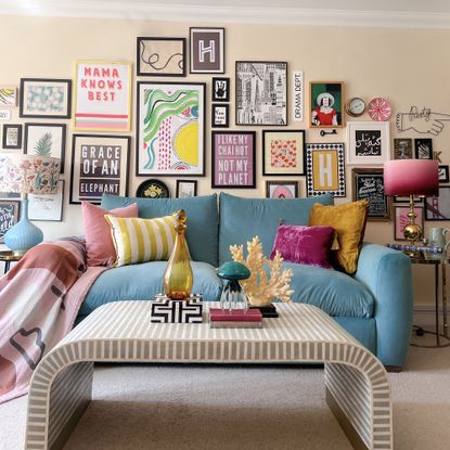Colourful wall art in black frames arranged to fill the wall in a soft yellow living room with a bright blue sofa and curved coffee table