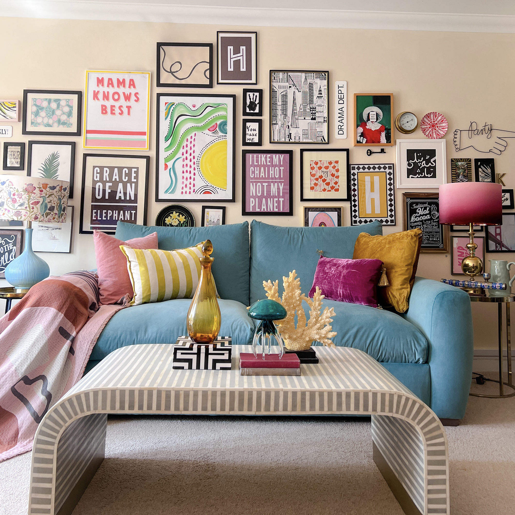 Colourful wall art in black frames arranged to fill the wall in a soft yellow living room with a bright blue sofa and curved coffee table