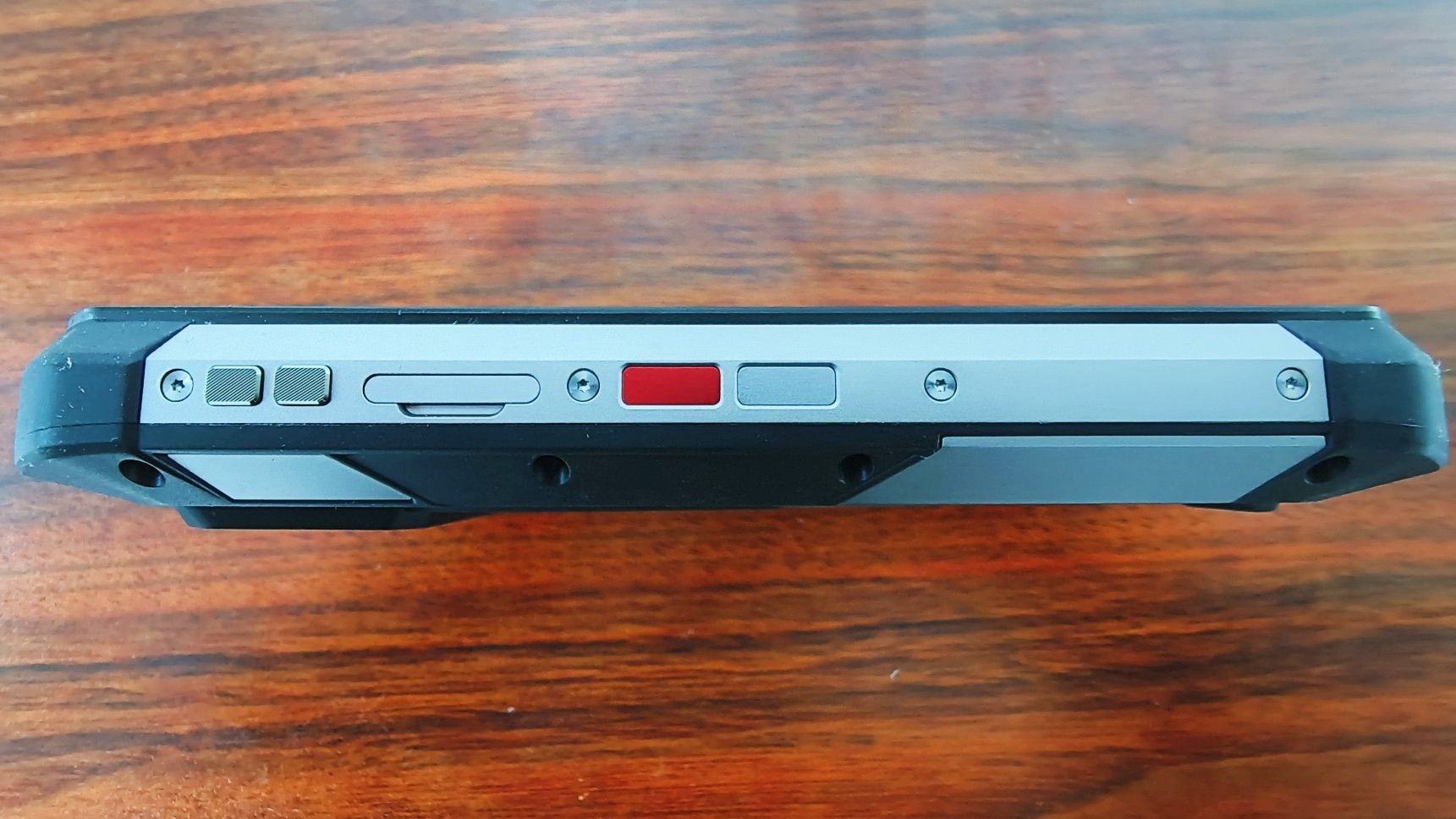 The left side of the rugged smartphone shows two silver buttons, a SIM tray slot, a prominent red button, and another gray button.