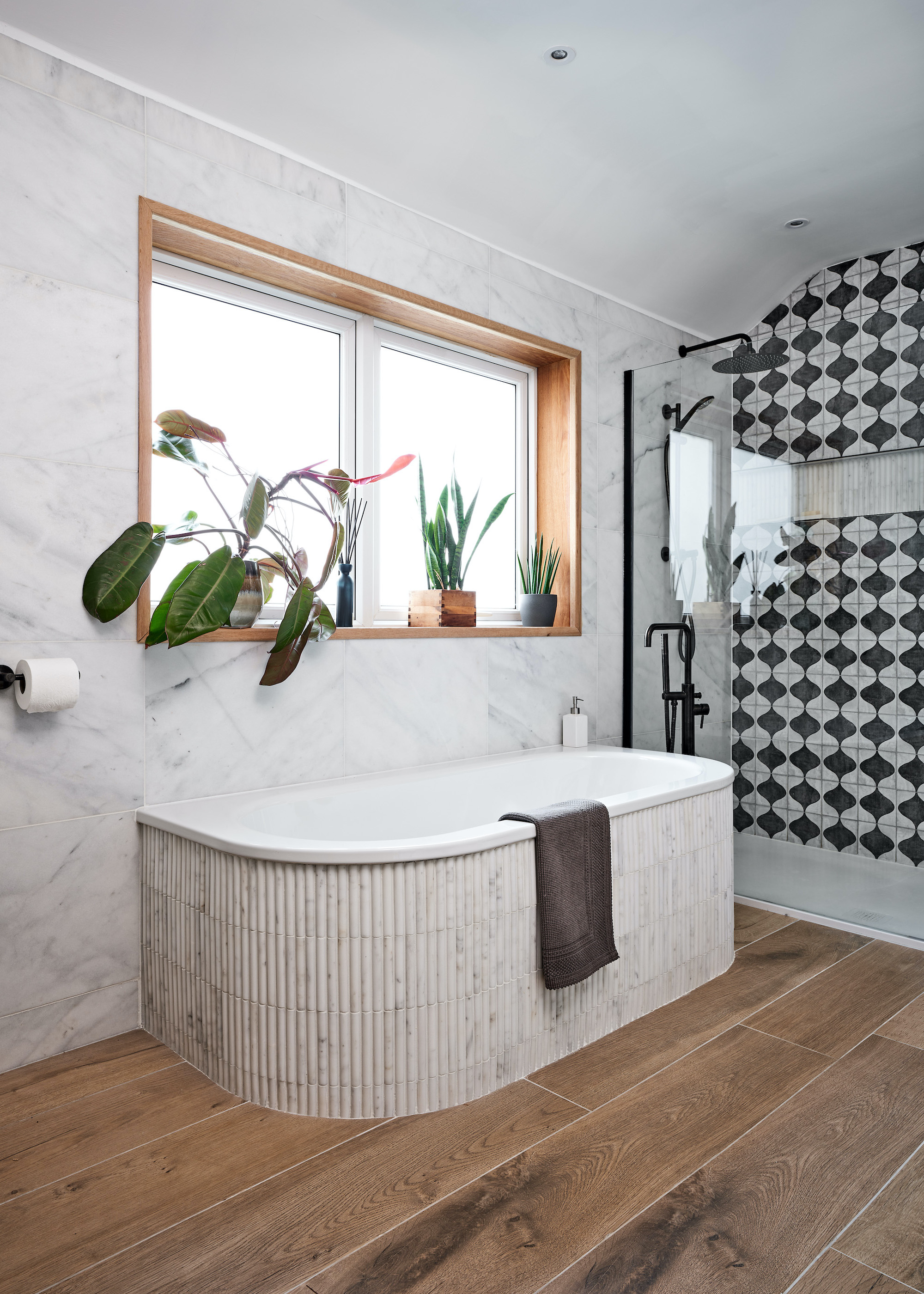 A curved bathtub with tiles around it as well as wall tiles around the bath tub and a window above it with lots of plants on it