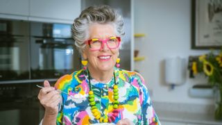 Prue in her kitchen smiling for the camera and wearing a bright top and necklace and holding a fork as if she is about to eat