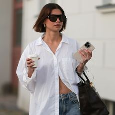 Sophia Geiss is seen wearing chunky silver hoop earrings, a silver nose ring, black aviator sunglasses from Vehla Eyewear, an oversized white structured shirt from Black Palms, wide-leg blue-grey Bermuda shorts from Drykorn, a large rectangular black leather “Birkin” bag from Hermès, a gold bangle, a minimalist silver ring, and black flip-flops from Havaianas, while holding a coffee cup and an Apple iPhone paired with Apple Wired EarPods during Berlin Fashion Week SS26 on June 30, 2025 in Berlin, Germany