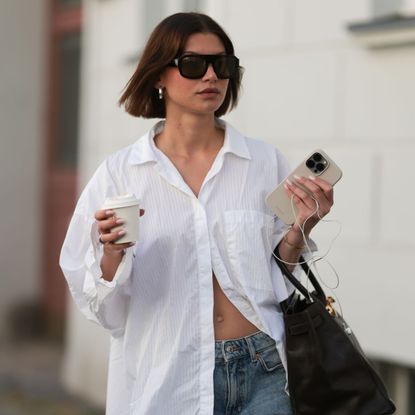 Sophia Geiss is seen wearing chunky silver hoop earrings, a silver nose ring, black aviator sunglasses from Vehla Eyewear, an oversized white structured shirt from Black Palms, wide-leg blue-grey Bermuda shorts from Drykorn, a large rectangular black leather &ldquo;Birkin&rdquo; bag from Herm&egrave;s, a gold bangle, a minimalist silver ring, and black flip-flops from Havaianas, while holding a coffee cup and an Apple iPhone paired with Apple Wired EarPods during Berlin Fashion Week SS26 on June 30, 2025 in Berlin, Germany
