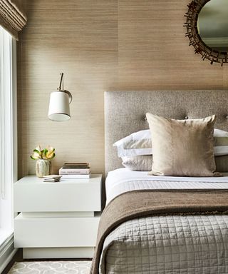 Modern bedroom with neutral brown and beige color scheme. Layered bedding, wall lamp above beside table.