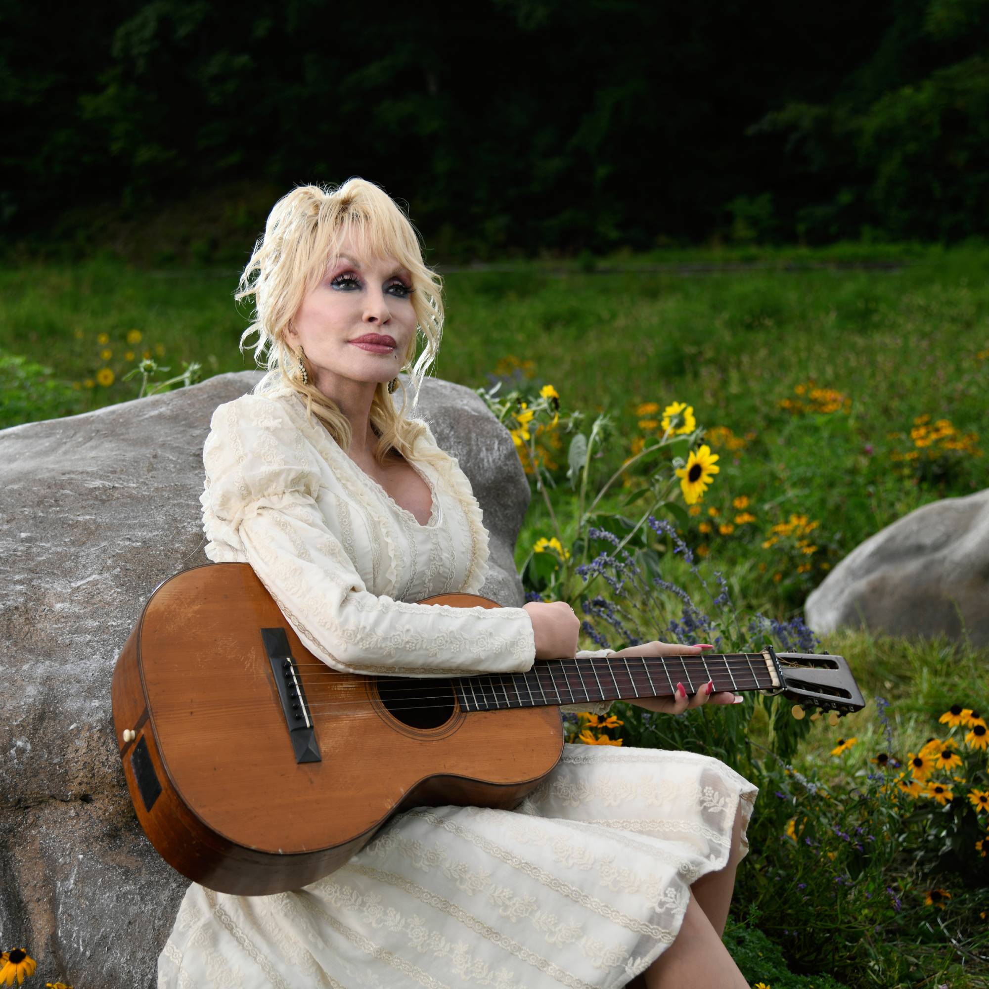 Dolly Parton looks into the distance holding a guitar in a field of flowers
