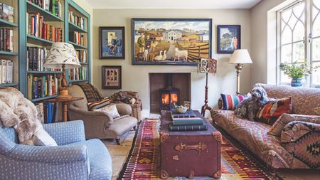 A cozy and eclectic living room with a library wall, lit wood burner, patterned large rug, vintage look trunk and a mismatched upholstered sofa and chairs.