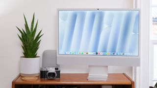 Apple iMac on a wooden desk against a white wall next to a lamp, camera and green plant