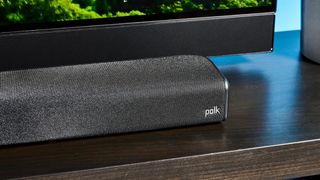 The Polk Audio Signa S4 being used in a lounge with a large TV. On the TV screen is a green forest scene. In the background is a blue wall.