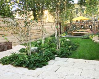 An urban backyard with white stone patio, lawn, and horizontal wood fence, and garden furniture at the bottom of the yard