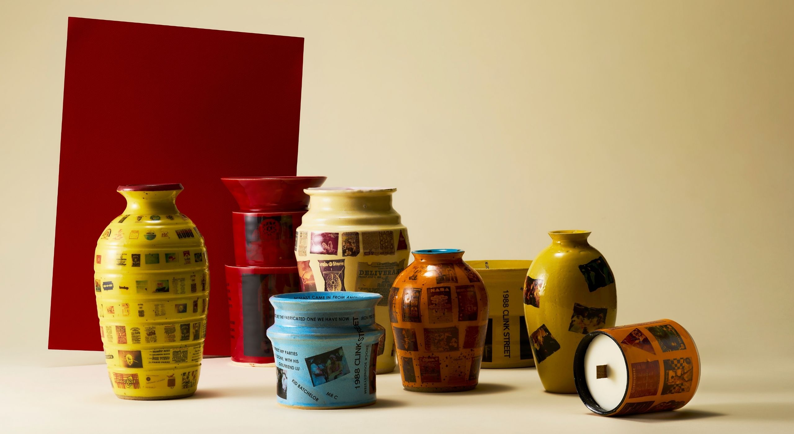 A series of ceramic vessels, decorated with overlaid newspapers and images excerpts glued onto their colorful surface, sit against a beige background and a red rectangle.