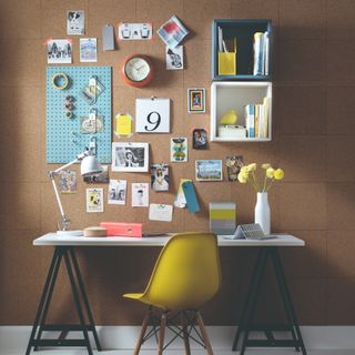 A home office with a white desk and a yellow chair and a whole wall covered in a cork board with pictures and notes stuck on top