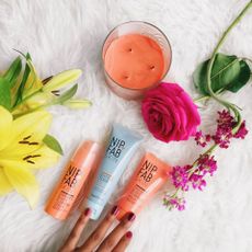 A hand touching Nip & Fab products next to pink rose, yellow lily, orange candle, on a white rug.