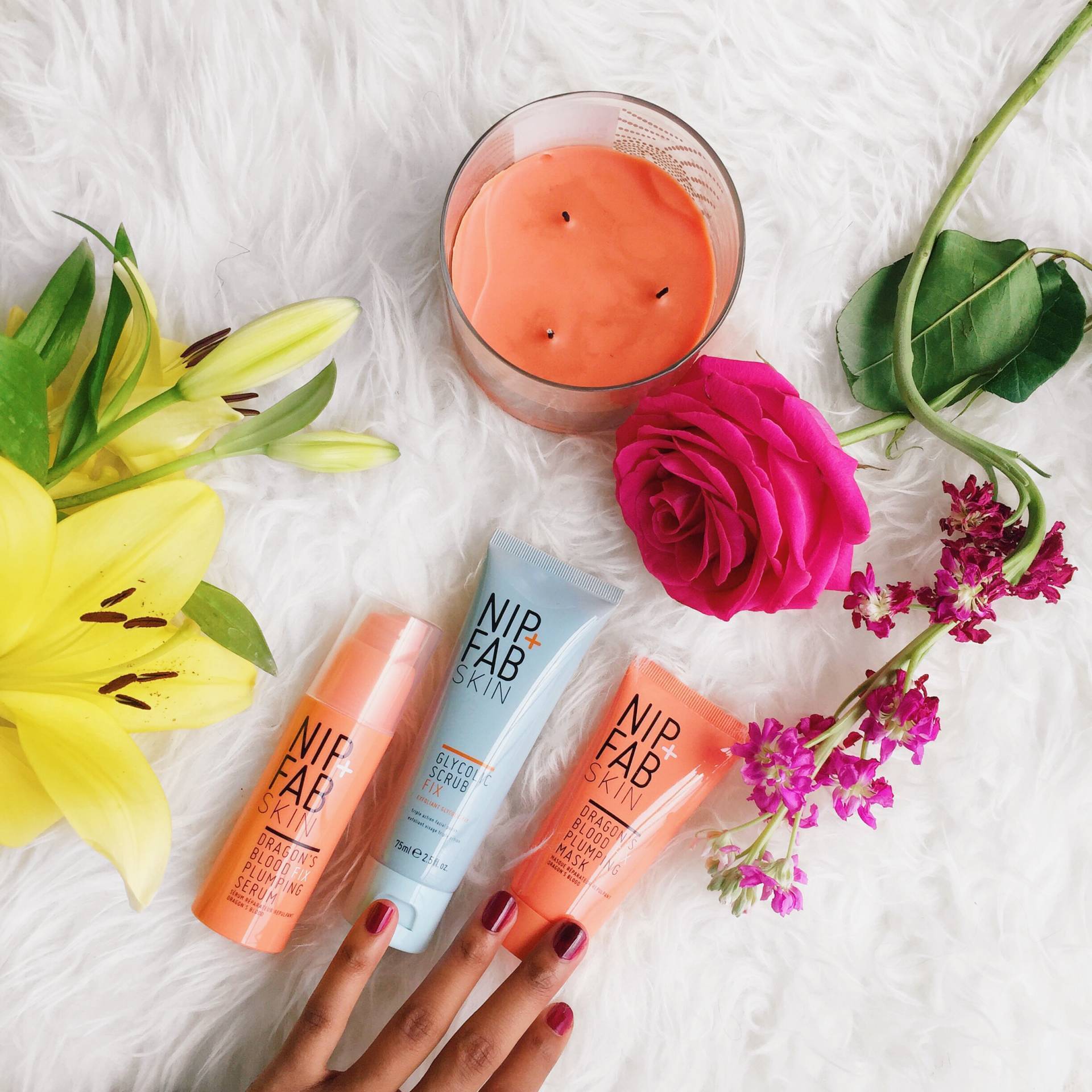 A hand touching Nip &amp; Fab products next to pink rose, yellow lily, orange candle, on a white rug.