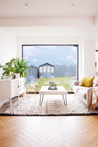 Part of a larger open-plan space, a living area with monochrome patterned Scandi-style rug, vintage-style white sideboard, sofa, and a large picture window
