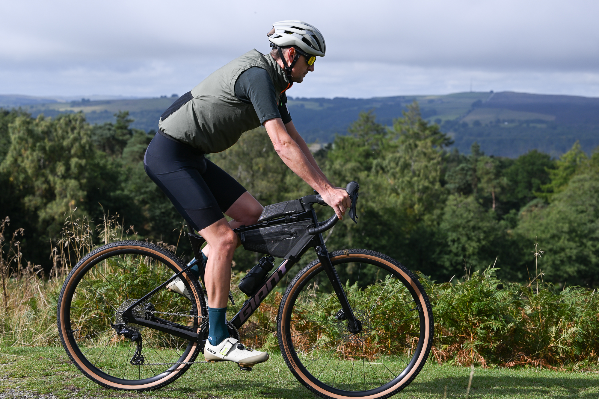 a male rider cycling a black gravel bike on a grassy trail wearing a green jersey, green gilet, black shorts and sunglasses
