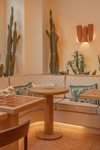 A dining room features wooden tables, textile banquettes and cushions with decorative patterns, tall cacti, and suffused lighting, all in the tones of terracotta.