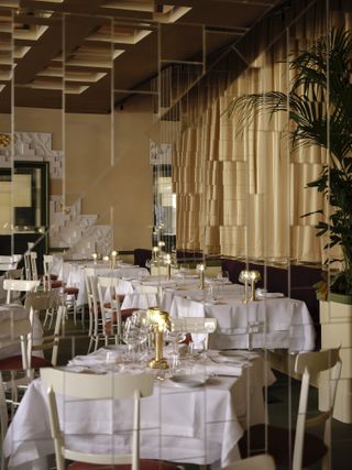 A 1970s-inspired dining room in a hotel features mirrored walls made of reflective, square tiles, linear white tables and chairs, and golden table lamps as well as plants.