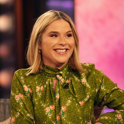 a white woman with short, blonde hair wearing a green, floral print, high neck blouse while being interviewed on the set of a talk show