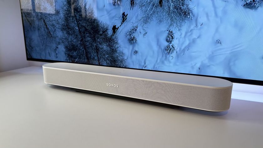 A white Sonos Beam Gen 2 soundbar photographed on a white desk in front of an OLED TV